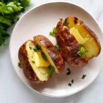 Air Fryer Baked Potatoes sit cut open on a white platter. They have pats of butter and fresh herbs on them.