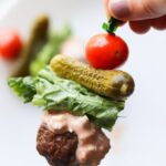 An Air Fryer Burger Bite on a toothpick with dressing, lettuce, a small pickle and a cherry tomato.