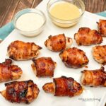 Air Fryer Chicken Bites wrapped in bacon and sitting on a white cutting board next to two glass bowls of dip.
