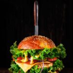 A single Air Fryer Turkey Burger with all the fixings. A knife sits upright in the middle of the burger. Cheese melts over the patties and lettuce in this Air Fryer Turkey Burgers Recipe.