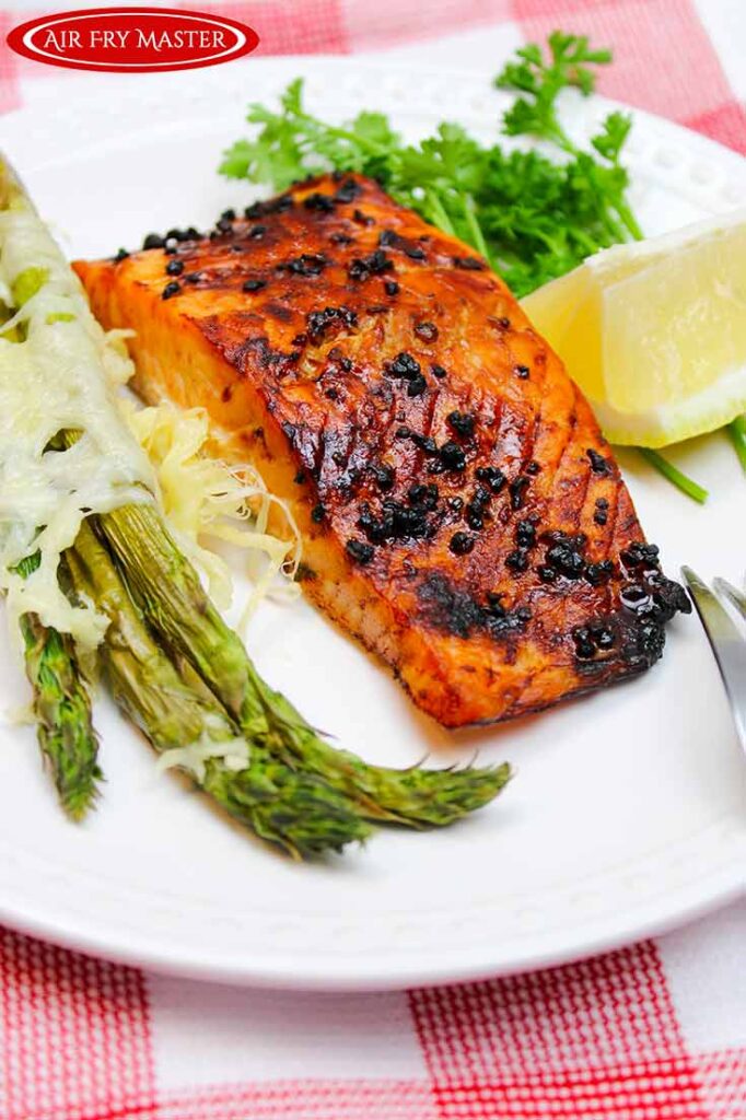 A front view of a white plate holding a Air Fryer Honey Glazed Salmon fillet, some asparagus, a lemon wedge and fresh parsley.