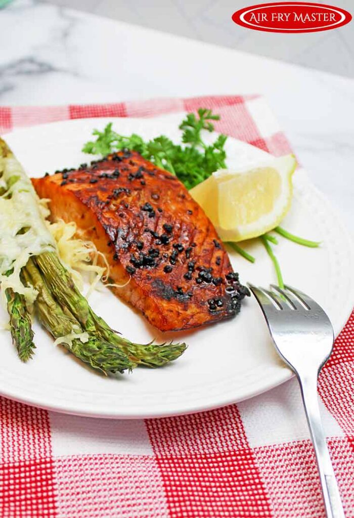 A side view of Air Fryer Honey Glazed Salmon served on a white plate. A serving of air fried asparagus sits on the plate to the left of the salmon. A lemon wedge and fresh parsley sit to the right.