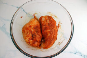 Two chicken breasts coated in spice mix and sitting in a mixing bowl.