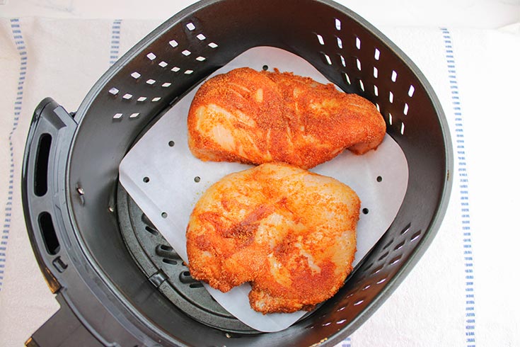 Two spice-coated chicken breasts sitting in a parchment-lined air fryer basket.
