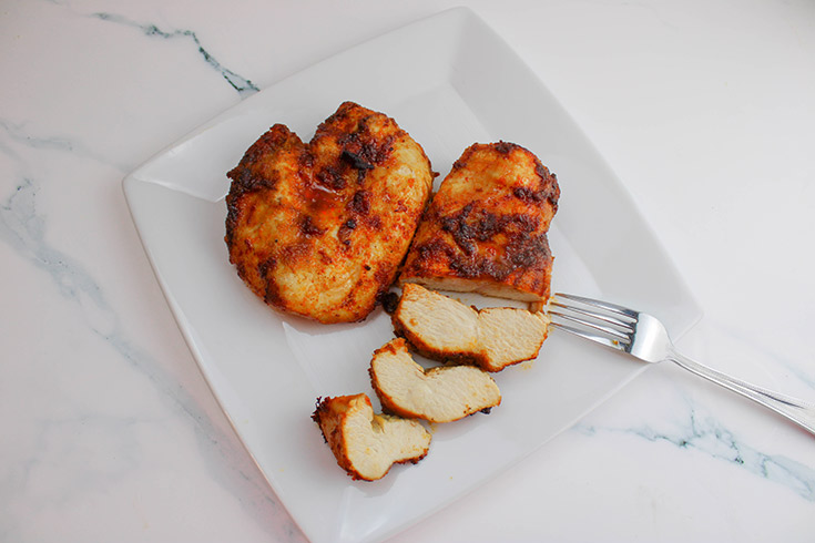 Two chicken breasts laying on a square, white plate. Once is halfway sliced with a fork laying next to it.