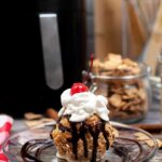 A finished Air Fried Ice Cream ball on a plate, topped with chocolate sauce, whipped cream and a cherry.